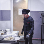 A professional chef in a black uniform and hat works at a stainless steel station, sprinkling toppings onto a pizza dough as bowls of ingredients and flour lie nearby in a busy kitchen.