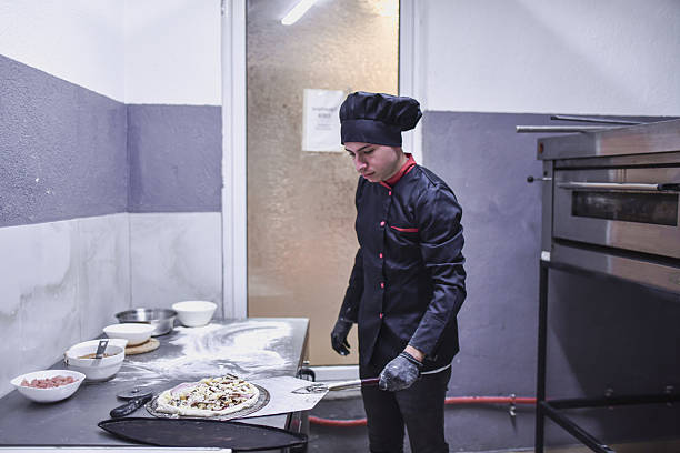 A professional chef in a black uniform and hat works at a stainless steel station, sprinkling toppings onto a pizza dough as bowls of ingredients and flour lie nearby in a busy kitchen.